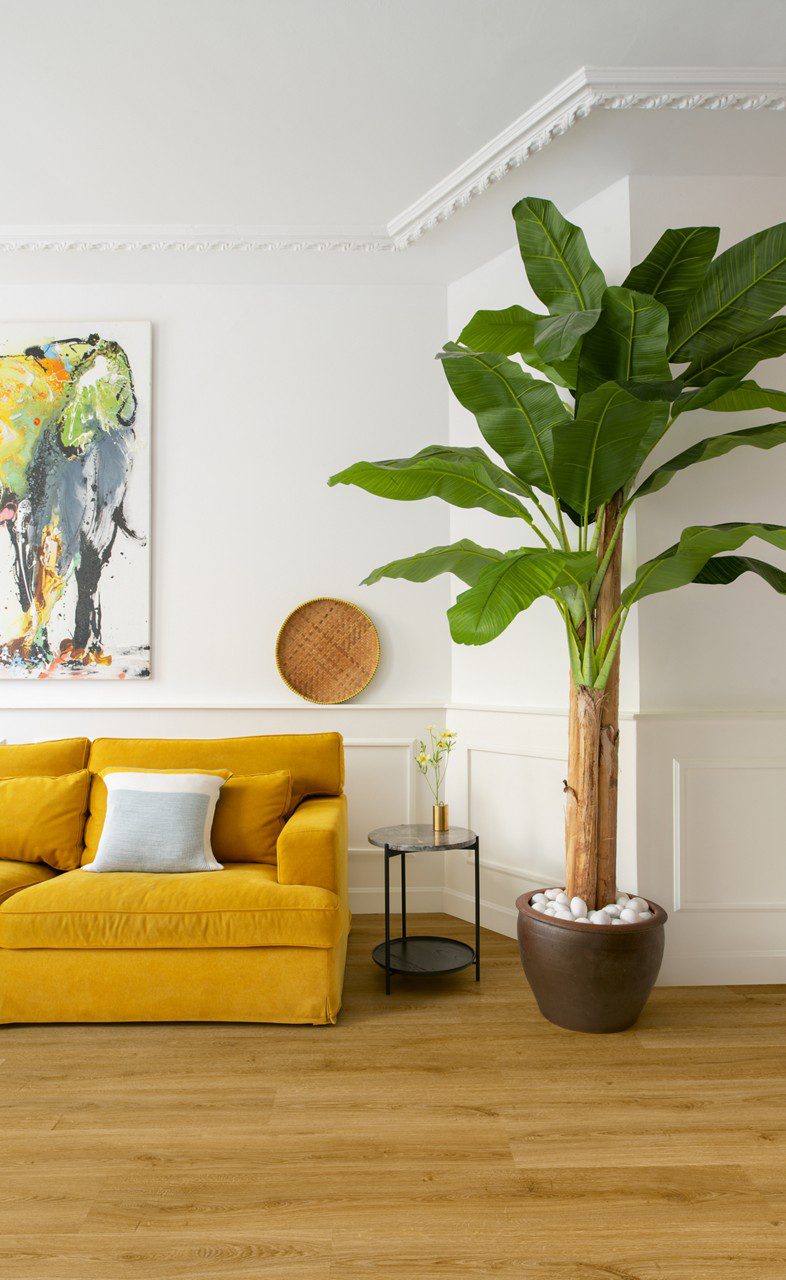 LVT parkett Quick-Step Bloom Botanic Smoked Oak paigaldatud elutuppa. Kollane diivan, suur palmipuu, maal seinal.