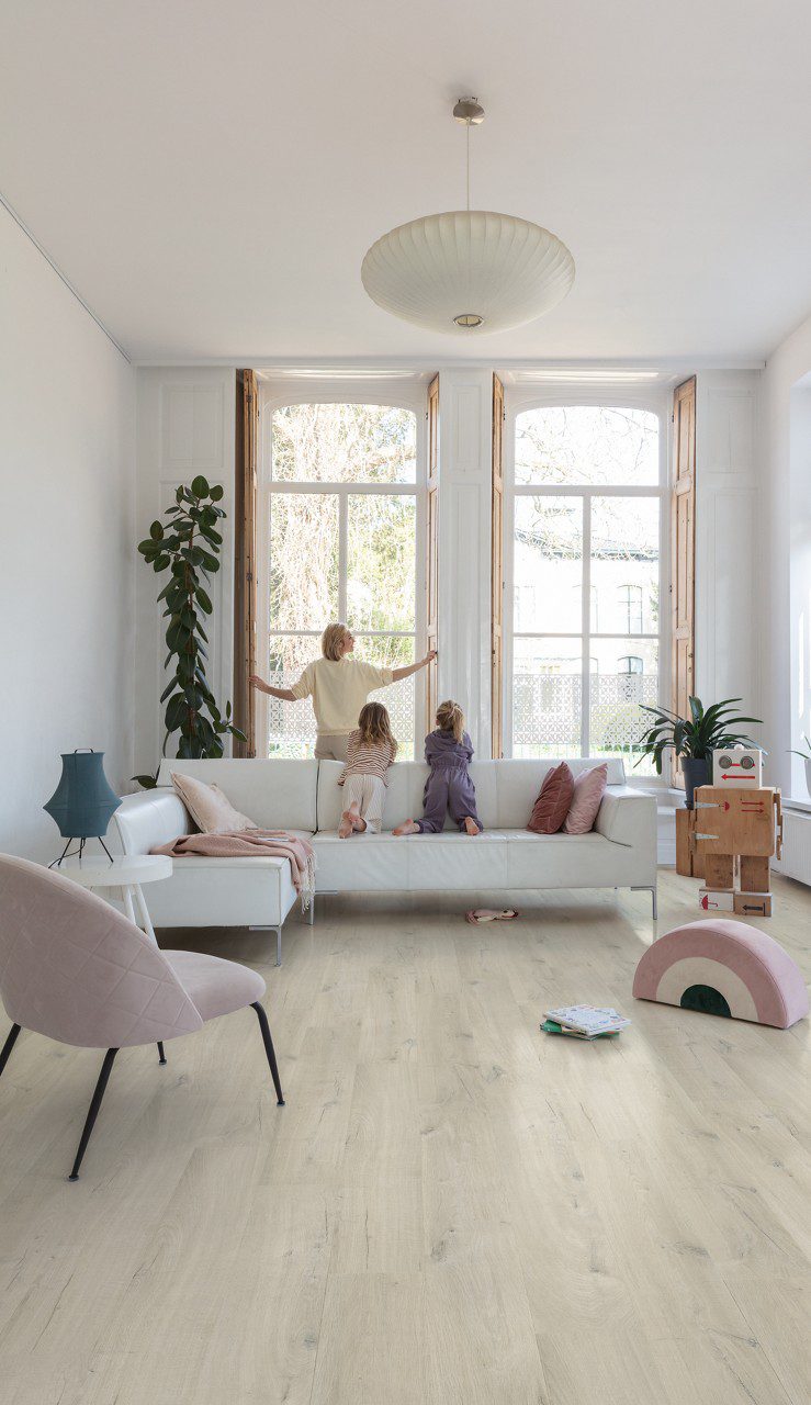 LVT parkett Quick-Step Bloom Cotton Oak White Blush paigaldatud elutoa põrandale. Ema sulgeb puidust aknakatteid ja lapsed vaatavad seda valgelt nahast diivanilt.
