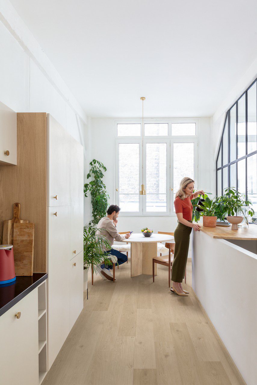 LVT parkett Quick-Step Blos coast tamm sand paigaldatud köögi põrandale. Naine kastab potilille töötasapinnal. Mees istub söögilaua taga ja vaatab telefoni.