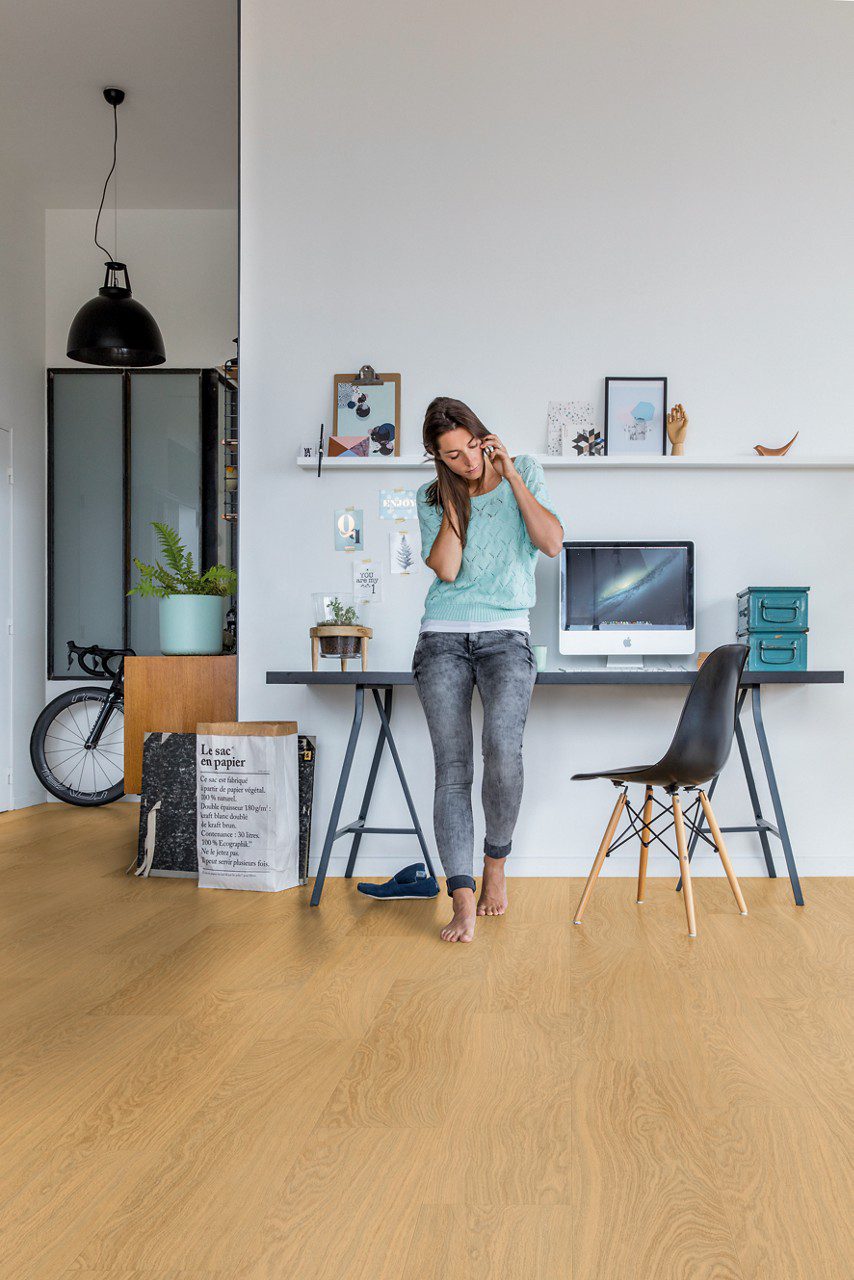 LVT Quick Step Bloom Pure Oak Honey paigaldatud kodukontori põrandale. Naine räägib telefoniga toetudes kontori lauale. Tool, jalgratas, apple arvuti.