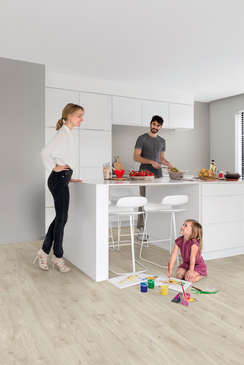 Vinüülparkett Quick-Step Blos Canyon tamm beige paigaldatud köögi põrandale. Valge köögimööbel ja köögisaar millel isa lõikab perele salatit. V'ike tütar joonistab õrandal värvidega pilte ja ema jälgib teda toetudes käega köögisaarele.