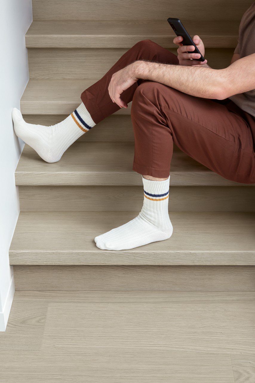 Vinüülparkett Quick-Step Blos Chia tamm helehalli tooniga paigaldatud koridori põrandale ja trepile. Mees istub trepil ja vaatab telefoni.