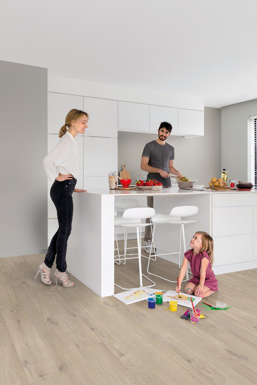 Vinyylilattia Quick Step Bloom Cotton Oak Beige asennettuna keittiön lattialle, vaalea puuvillabeige tammen sävy ja lankkudesign, isä laittaa ruokaa ja äiti katsoo lasta, joka piirtää kuvia lattialla.
