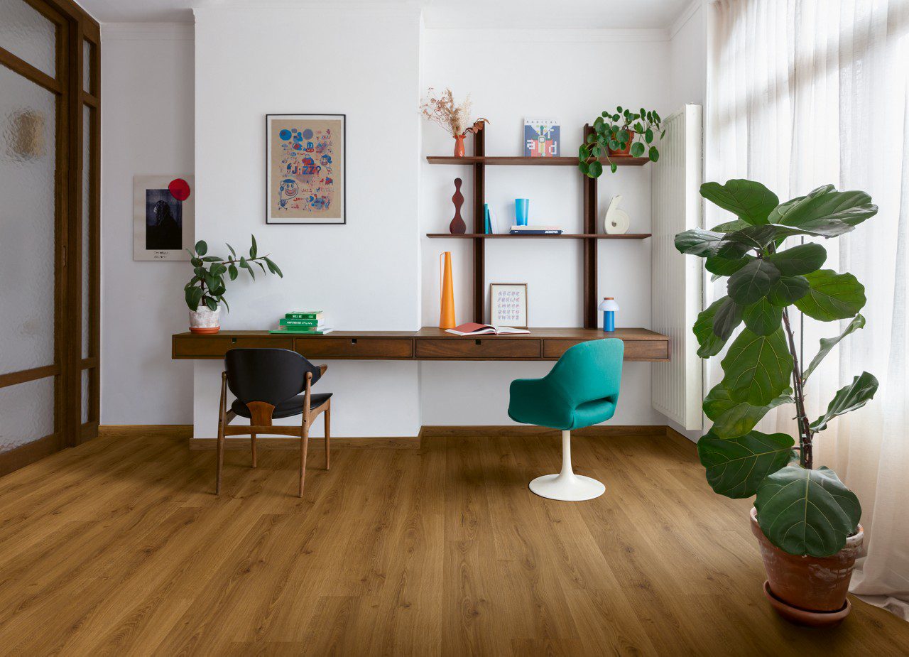 Виниловый ламинат Quick Step Bloom Botanic Caramel Oak уложен на полу в домашнем офисе, письменный стол, светло-голубое офисное кресло, пальма и цветы.