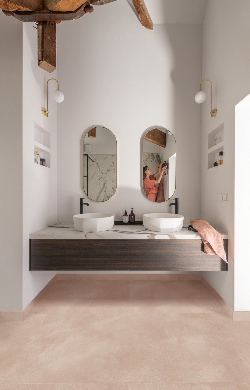 LVT Quick-Step Illume Soft blush paigaldatud vannitoa põrandale. Topelt vannitoamööbel valgest marmorist tasapinnaga millel kaks stiilset tasapinnapealset valamut. Seinapeeglist paistab naisterahvas.