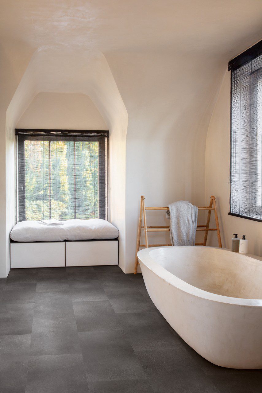 Vinüülparkett Quick-Step Oro Volcanic rock tumehalli tooni ja faasitud looduskivi disainiga. Paigaldatud vannituppa, suur looduskivi vann. Kapp akna all millel pehmendus istumiseks.