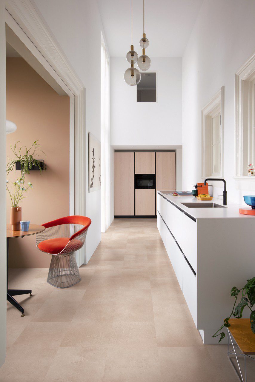 Vinyl floor tiles Quick-Step Oro Coral Rock installed on kitchen floor. White and beige kitchen furniture. Kitchen table and red stool.