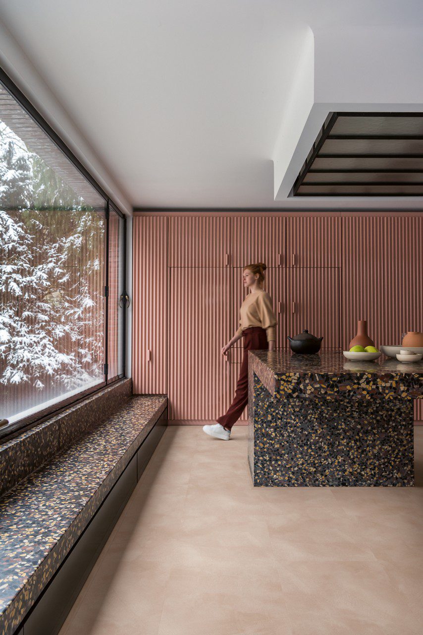 Vinylboden in Betonoptik Quick-Step Illume Soft Blush, verlegt auf dem Küchenboden – rosafarbene, in die Wand integrierte Küchenschränke und eine große Kücheninsel in Steinoptik; heller, zart rosébeiger Ton mit dezentem, steinartigem Design.