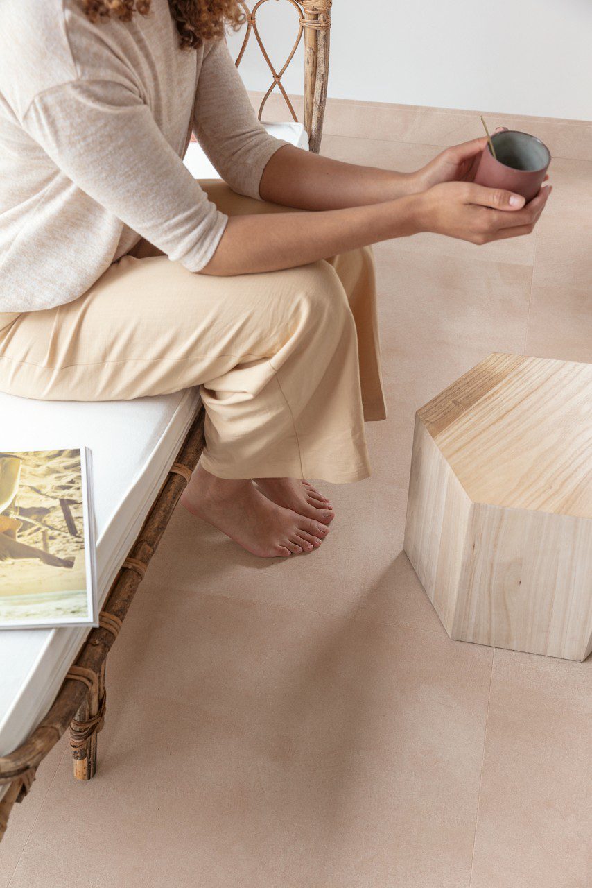 Vinylboden in Betonoptik Quick-Step Illume Soft Blush, verlegt auf dem Wohnzimmerboden – eine Frau sitzt auf dem Sofa und hält eine Tasse Tee in der Hand; heller, zart rosébeiger Ton mit dezentem, steinartigem Design.