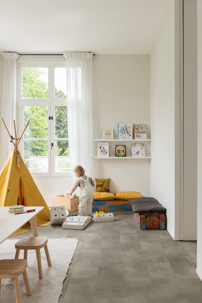 Vinylboden in Steinoptik Quick-Step Oro Concrete Rock, verlegt auf dem Boden eines Kinderzimmers – gelbes Tipi-Zelt im Indianerstil, Spielzeugkiste, Sitzkissen sowie Tisch mit Stühlen; ein Kind nimmt ein Spielzeug aus der Kiste.