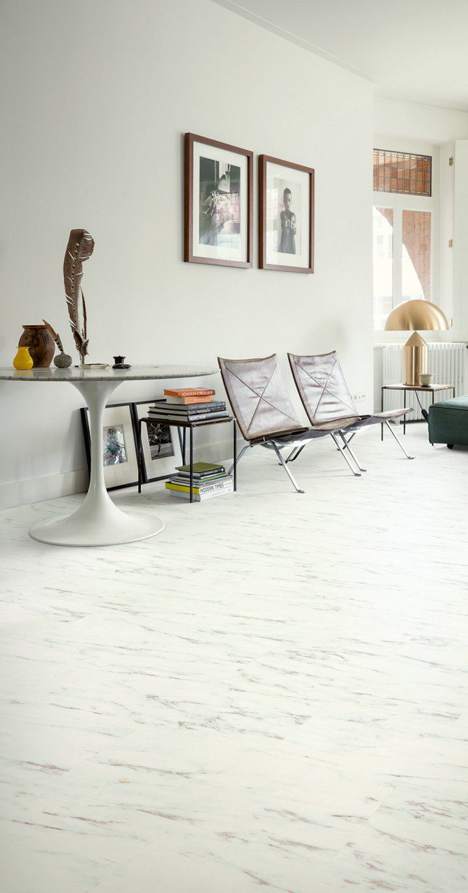 Vinylboden Stein Quick Step Oro Marble Carrara White im Wohnzimmer verlegt heller Carrara Marmor Look in Weiß mit feiner grauer Maserung braune Stühle mit Lederbezug runder Tisch und eckiger Tisch mit goldener Tischleuchte