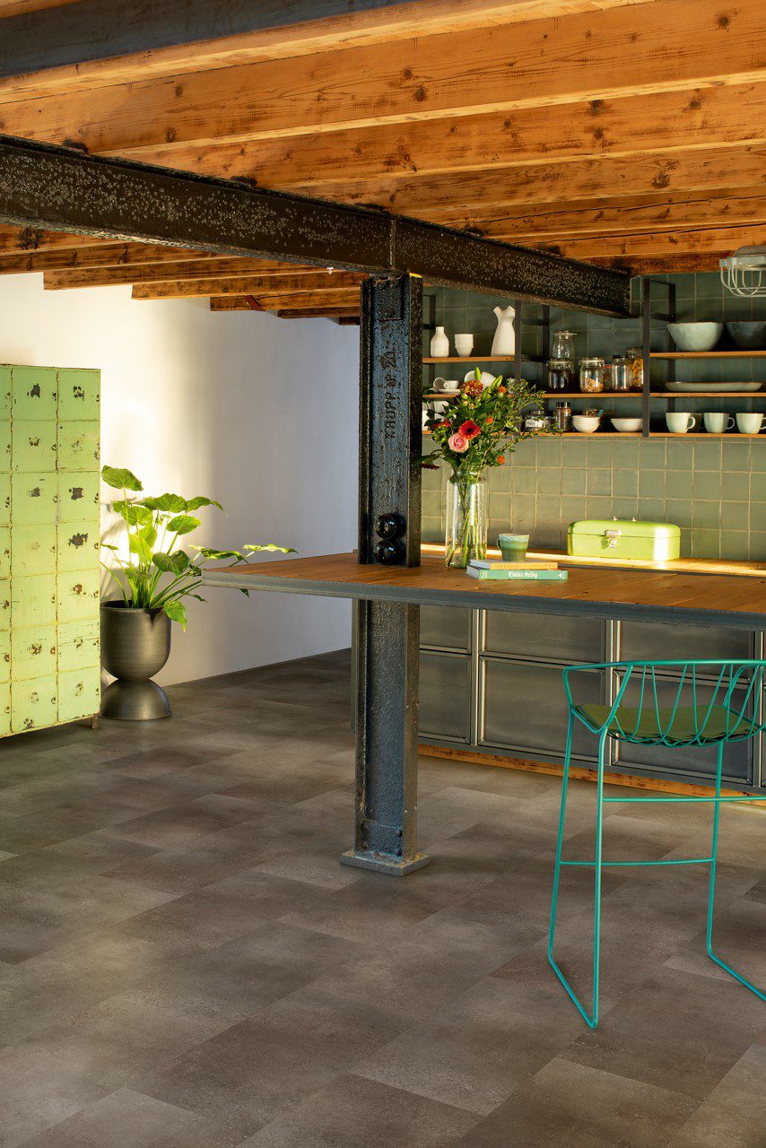 Vinylboden in Steinoptik Quick Step Oro Oxidized Rock in einer rustikalen Küche verlegt rostig kupferbrauner Farbton mit lebendiger Struktur industrieller Steinlook metallische alte Träger und Holzbalken an der Decke alter Küchenschrank Regale Esstisch und Stuhl