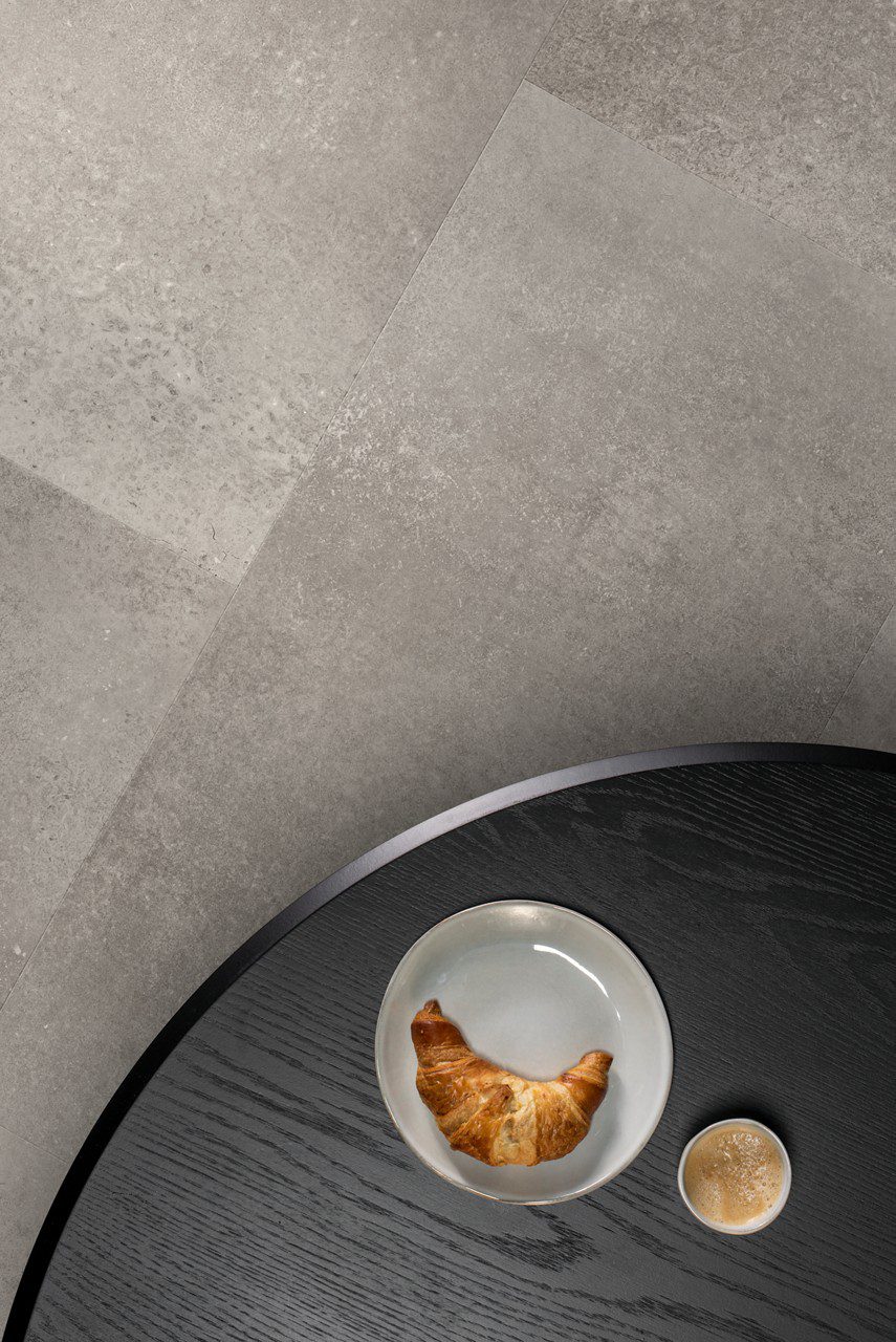 Кварц-винил Quick-Step Oro Concrete Rock уложен на полу кухни — кухонный стол, на котором лежат круассан и чашка кофе.