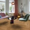 Helles, stilvolles Wohnzimmer mit Fischgrätparkett aus Eiche Mix (140 mm, geölt). Die Einrichtung kombiniert zartrosa Sofa, salbeigrünen Designstuhl und einen modernen Couchtisch mit goldenen Akzenten. Große weiße Flügeltüren und Fenster bringen viel Tageslicht in den Raum. Warme Vorhänge, moderne Kunstobjekte und eine elegante Ambienteleuchte schaffen eine harmonische und einladende Atmosphäre. Fischgrätparkett Eiche Mix geölt & gewachst 140 mm – 9