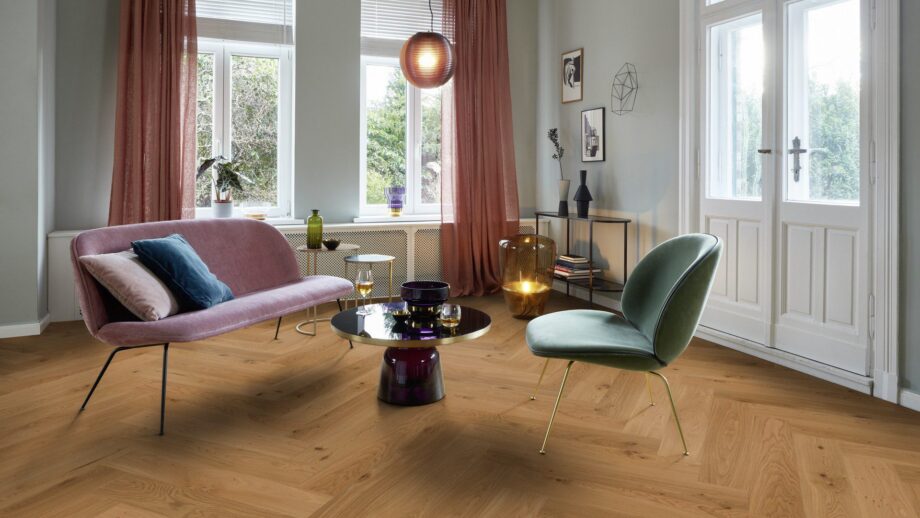 Helles, stilvolles Wohnzimmer mit Fischgrätparkett aus Eiche Mix (140 mm, geölt). Die Einrichtung kombiniert zartrosa Sofa, salbeigrünen Designstuhl und einen modernen Couchtisch mit goldenen Akzenten. Große weiße Flügeltüren und Fenster bringen viel Tageslicht in den Raum. Warme Vorhänge, moderne Kunstobjekte und eine elegante Ambienteleuchte schaffen eine harmonische und einladende Atmosphäre. Fischgrätparkett Eiche Mix geölt & gewachst 140 mm – 9
