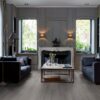 LVT parkett Quick-Step Blos Silk tamm dark grey paigaldatud klassikalise disainiga elutoa põrandale. Kaks diivanit suure diivanilaua ümber millel lilled ja küünland. Klassikalises stiilis valgest kivist kamin.