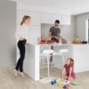 vinuulparkett-quick-step-blos-canyon-tamm-beige Vinüülparkett Quick-Step Blos Canyon tamm beige paigaldatud köögi põrandale. Valge köögimööbel ja köögisaar millel isa lõikab perele salatit. V'ike tütar joonistab õrandal värvidega pilte ja ema jälgib teda toetudes käega köögisaarele.