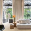 vinyylilattia-quick-step-blos-buttermilk-vaalea-tammi Vinyylilattia Quick Step Blos Buttermilk Vaalea Tammi asennettuna olohuoneen lattialle, vaalea kermaisen sävyinen tammen väri ja lankkudesign, kaunis sohva ja kukkia, mies astuu terassilta olohuoneeseen parvekeoven kautta.