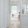 Vinyylilattia Quick Step Blos Vaalea Canyon Tammi Saw Cuts asennettuna makuuhuoneen ja kylpyhuoneen lattialle, lasiovi avoinna kylpyhuoneeseen, vaahtokylpy jossa yksi lapsi leikkii ja toinen seisoo lattialla vaahtoisin jaloin.